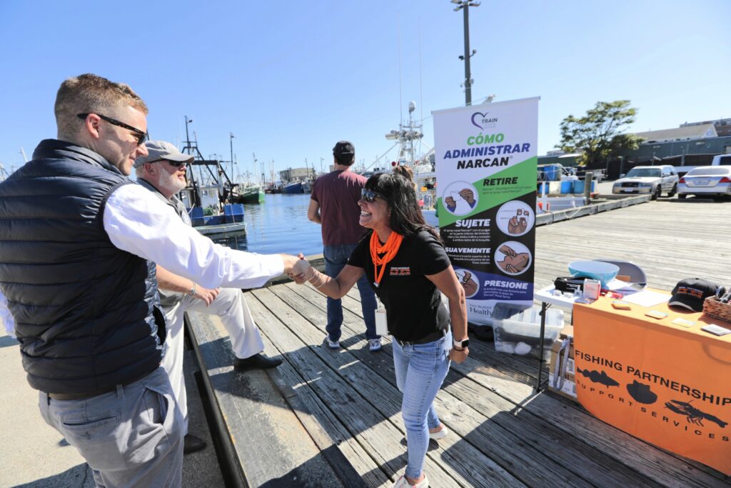 Woman with dark hair in a black t-shirt and jeans shakes hands with a man in a black vest while standing on a dock in front of an 'administer narcan' sign.